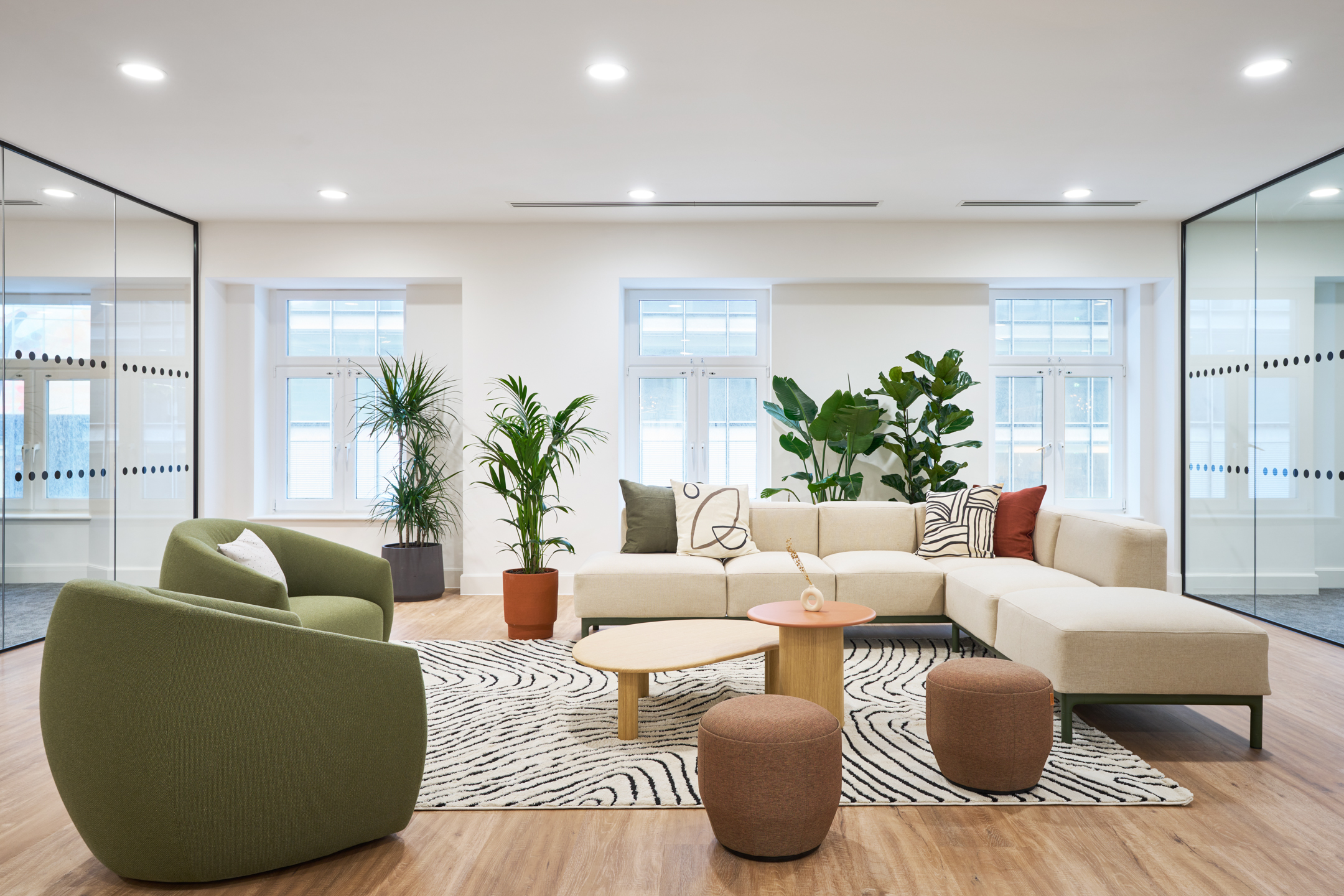 A bright lounge area at 7 Bishopsgate Level 4, with a large cream sofa, green accent chairs, and indoor plants against glass partition walls.