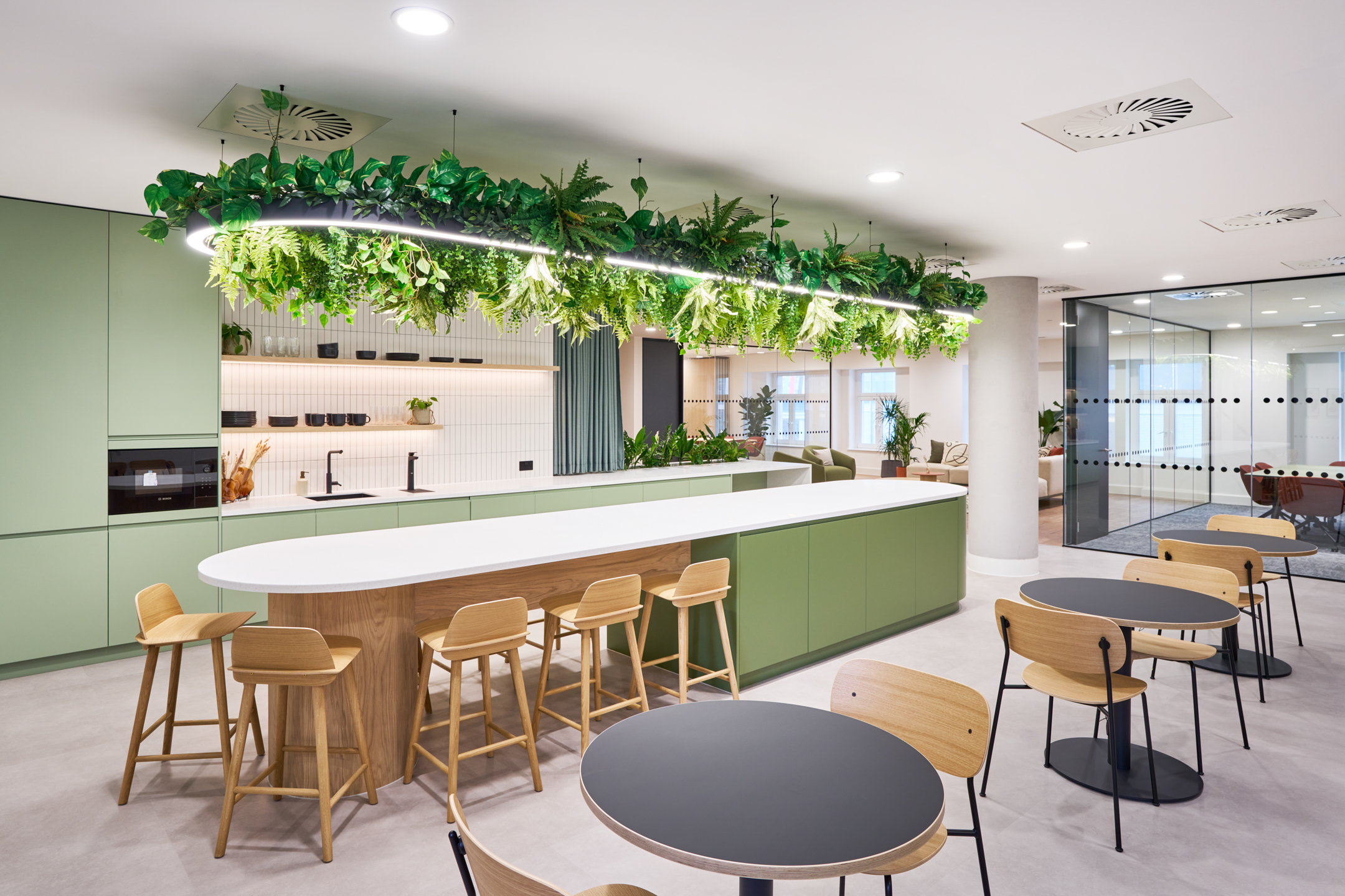 Contemporary teapoint area at 7 Bishopsgate Level 4, with green cabinets, wooden stools, a large island, and a ceiling adorned with hanging plants.