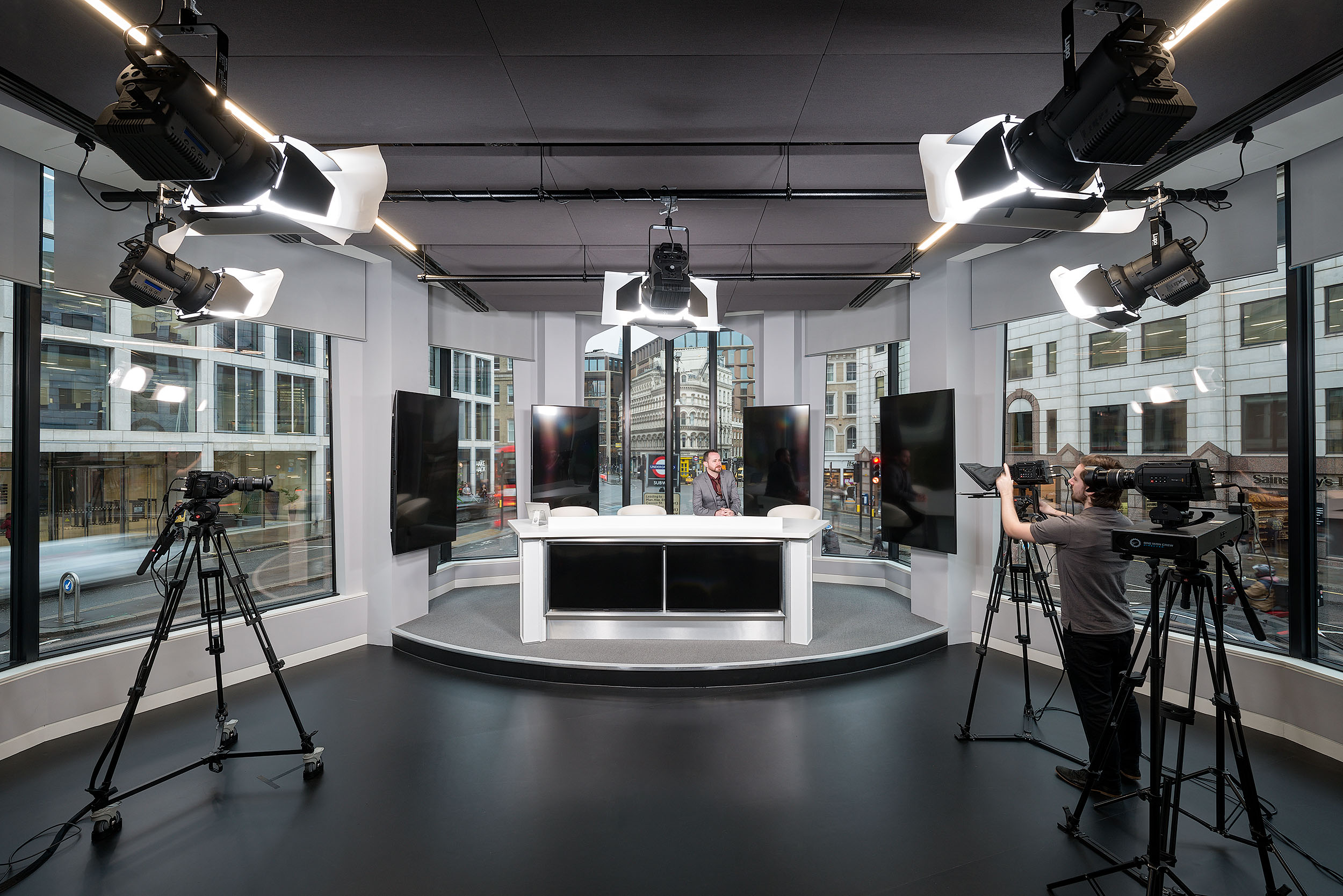 A modern studio setup at Asset TV, featuring multiple cameras, bright studio lighting, and large windows overlooking the street. The studio includes a central desk and screens positioned around the room.