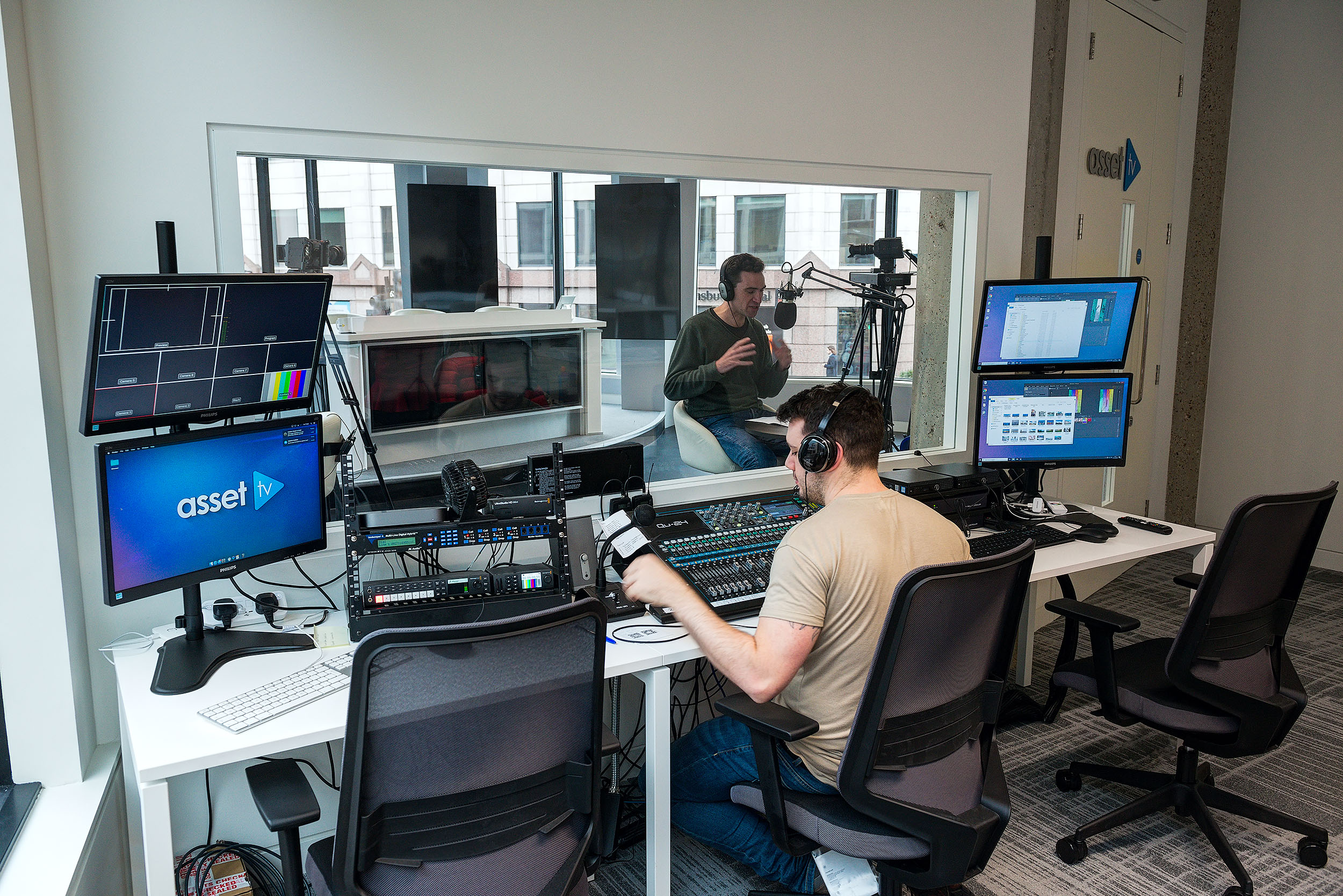 A view of the control room at Asset TV with a sound engineer operating equipment. Monitors display various audio and video feeds. A glass window separates the control room from the studio.