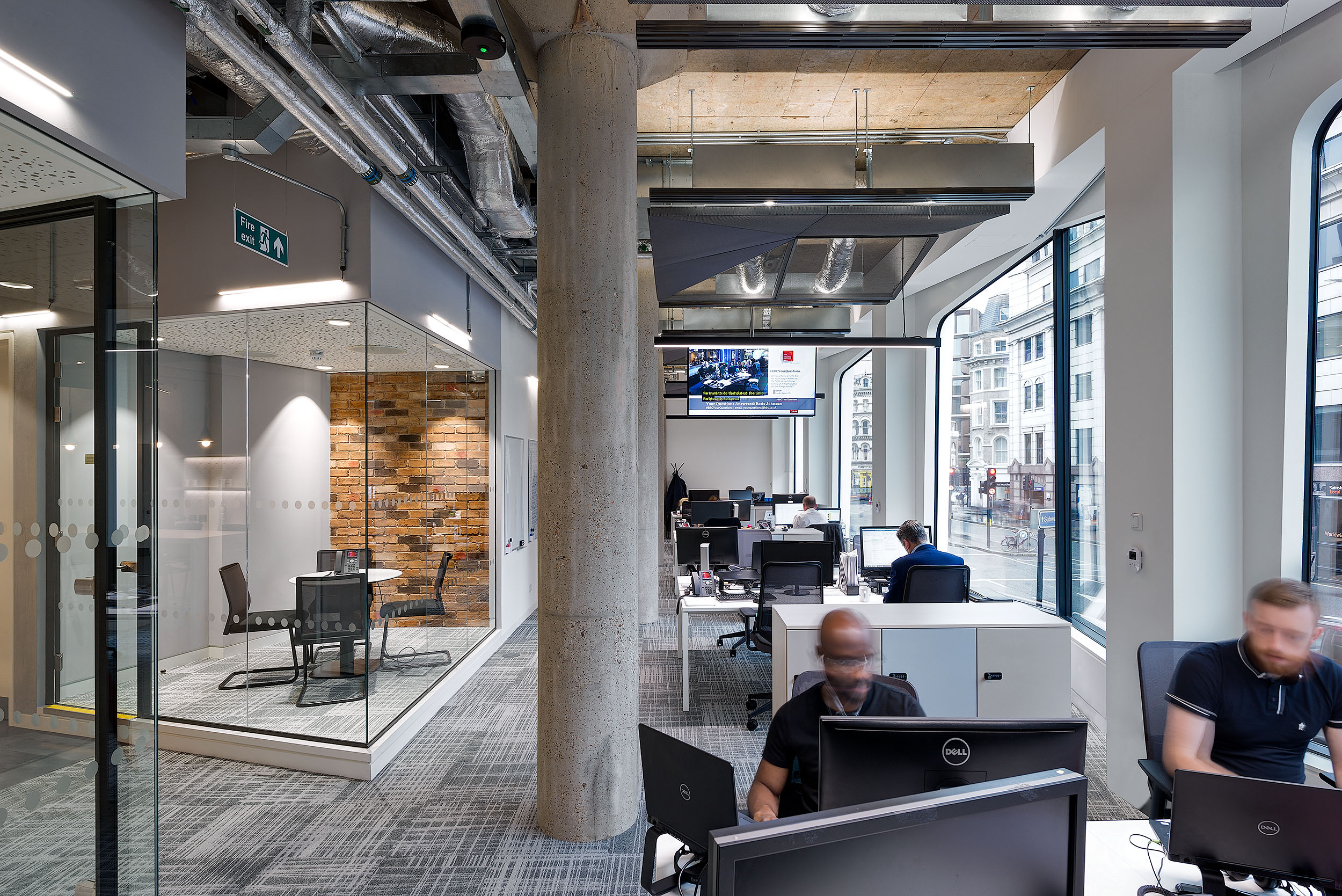 The open office area at Asset TV with staff working at desks, exposed ceiling with industrial-style ductwork, and floor-to-ceiling windows offering views of the surrounding buildings.