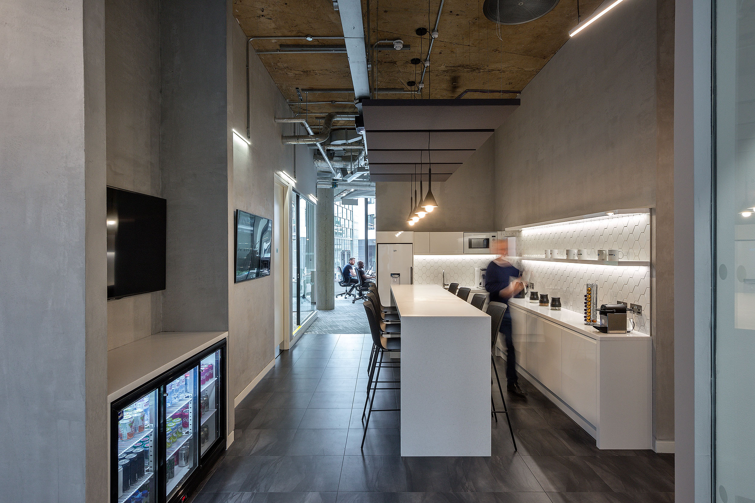 The teapoint at Asset TV with a modern design, featuring a long bar counter, hanging lights, and a beverage fridge. The space provides a casual area for employees.