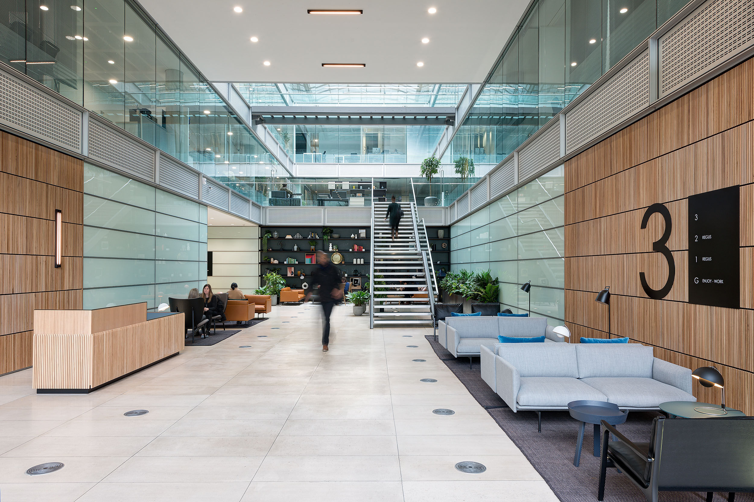 Reception area at Chiswick Business Park Building 3, with contemporary furnishings, a welcoming seating area, wooden panelled walls, and a central staircase leading to upper levels, all within a bright, glass-lined atrium.