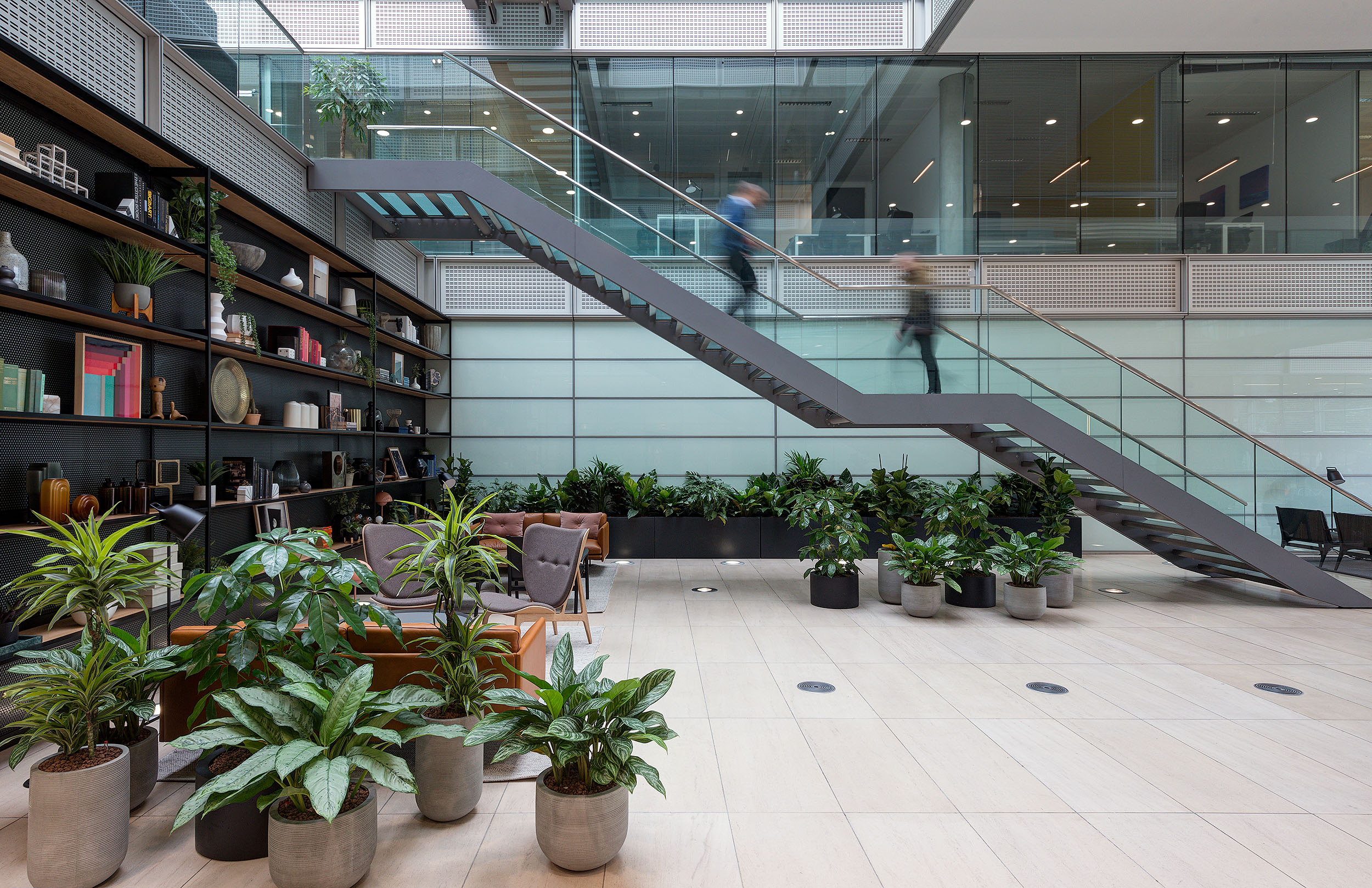 The reception atrium space in Chiswick Business Park Building 3, with a central staircase, abundant greenery, curated shelving with decorative items, and comfortable seating areas, creating a welcoming environment within a light-filled, glass-enclosed setting.