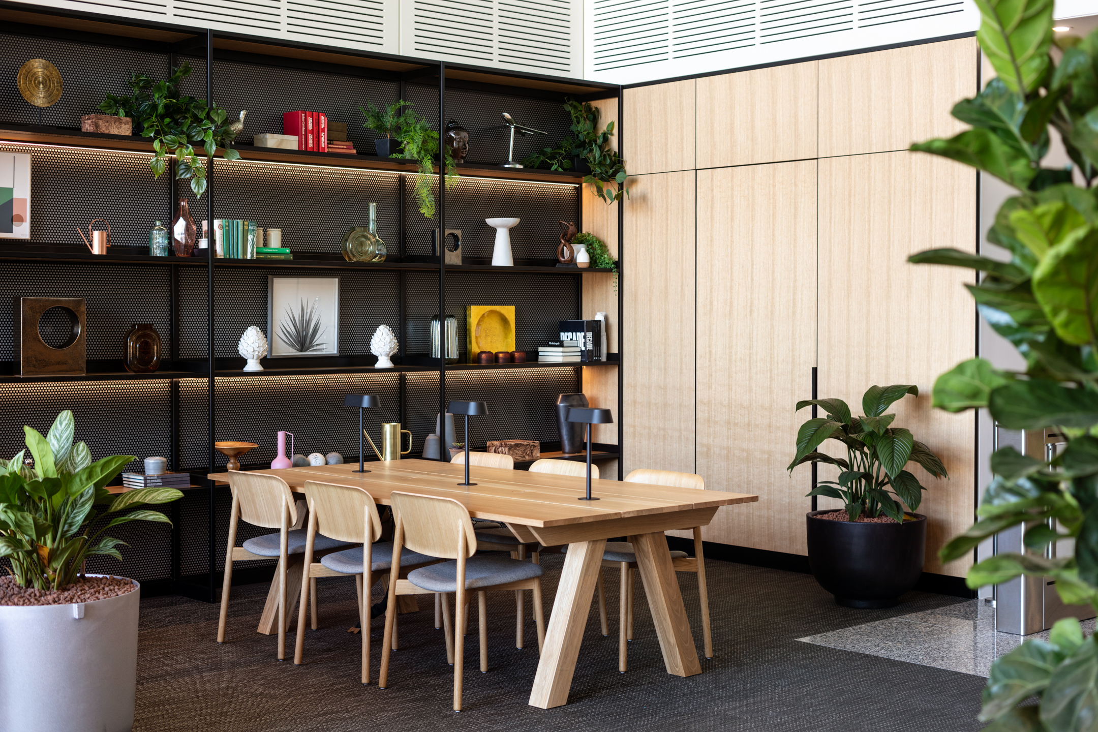 An elegant workspace featuring a large wooden table and stylish shelving units filled with plants and modern decorative items.