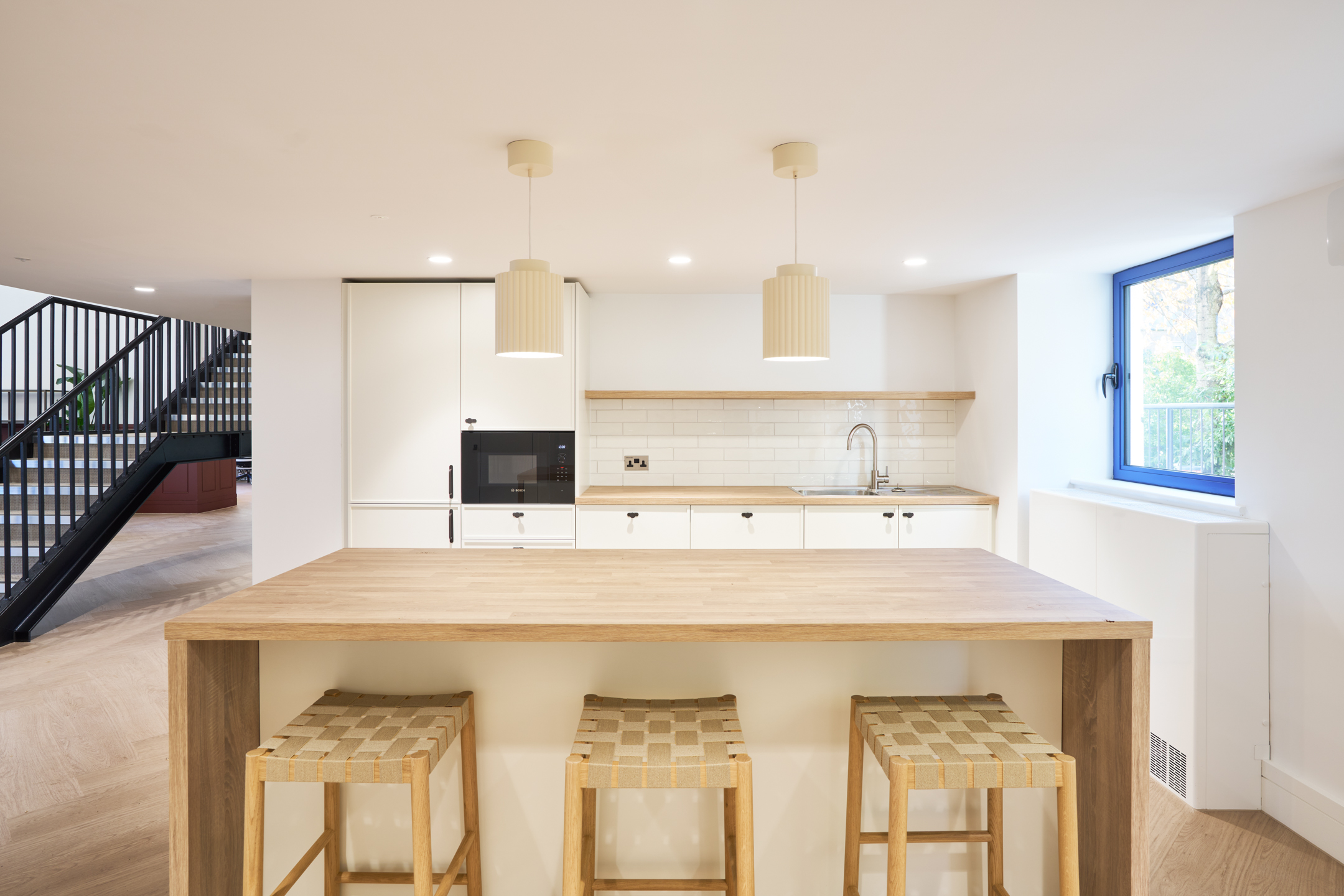 Minimalist kitchen area at 163-203 Eversholt Street, with a wooden island, bar stools, and pendant lights, in a bright, open space with a window.