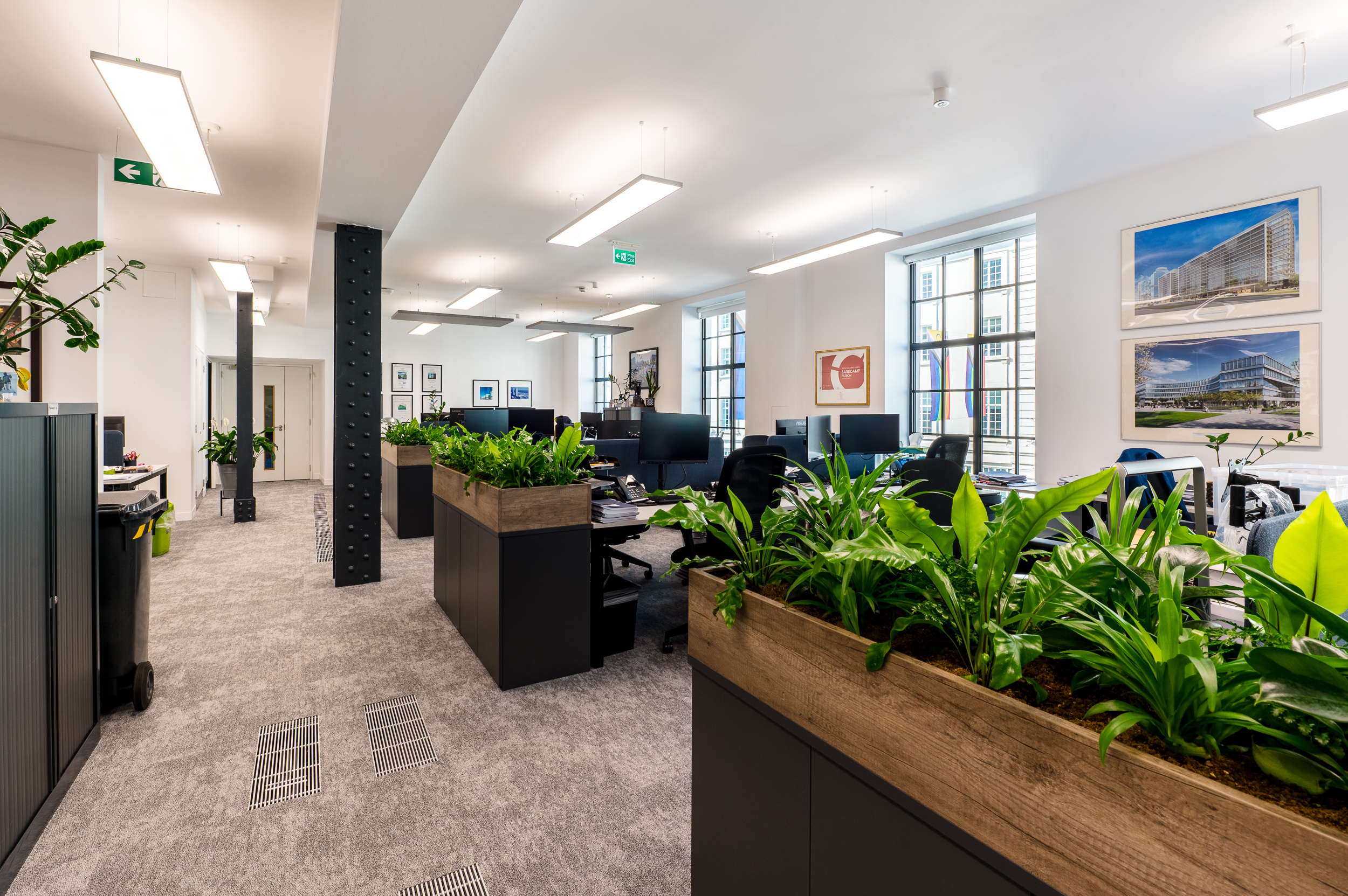 Open-plan office space with workstations, plants integrated into the design, and large industrial windows allowing natural light.