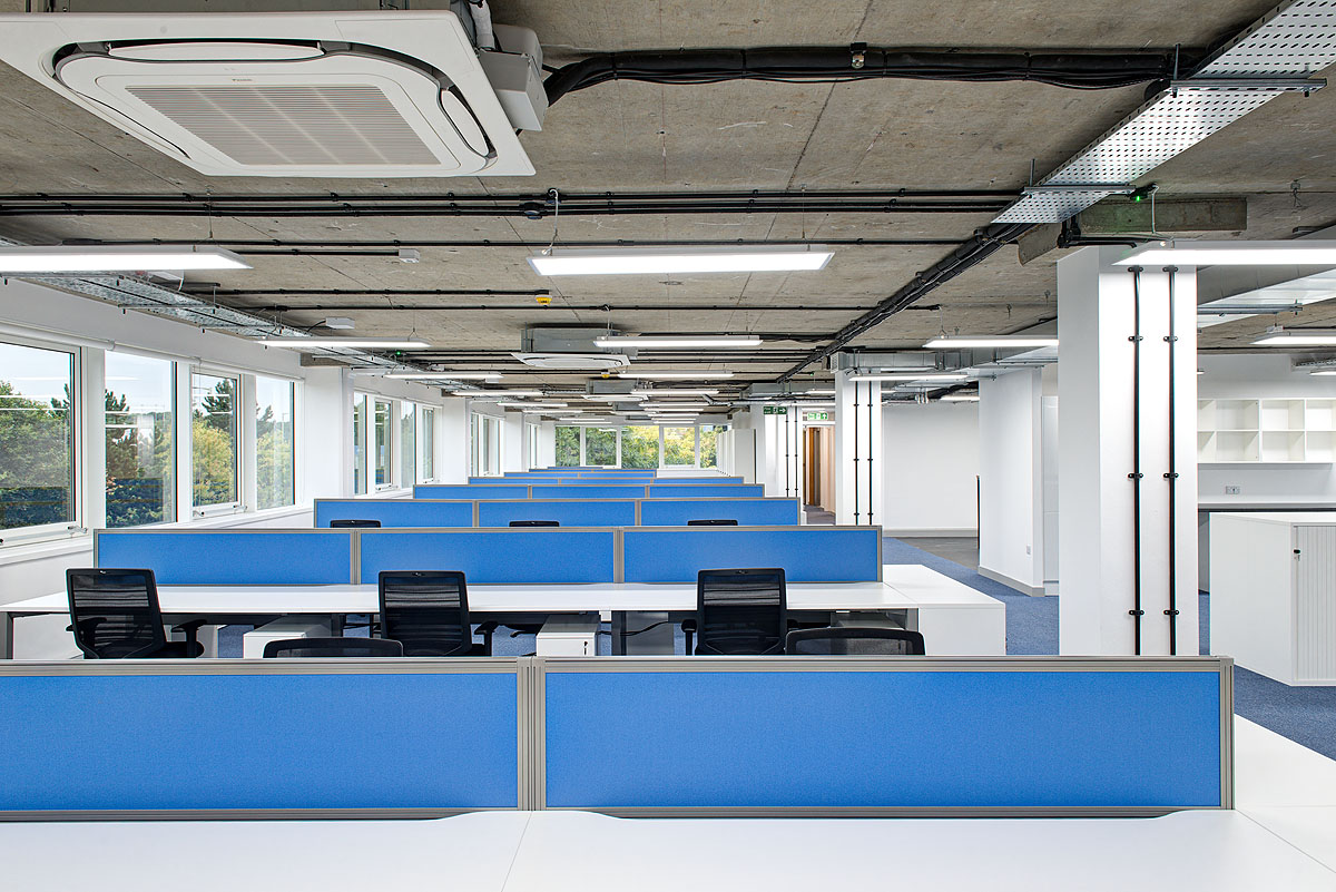 Open-plan workspace at Smiths Detection, featuring rows of blue-partitioned desks, large windows for ample natural light, and an industrial-style ceiling, fostering a spacious and productive office environment.