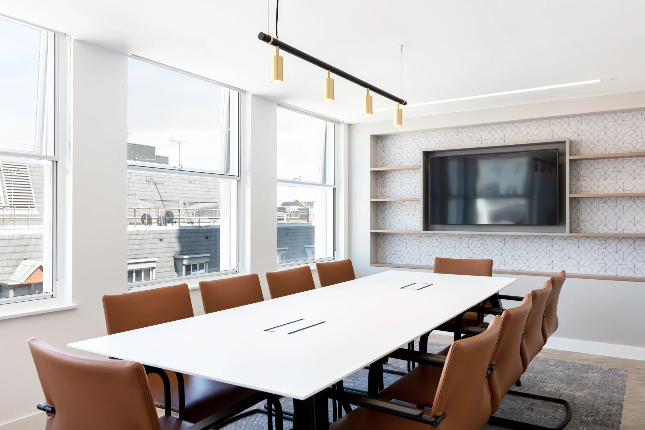 A bright conference room with a long white table, brown leather chairs, and a large wall-mounted TV screen.