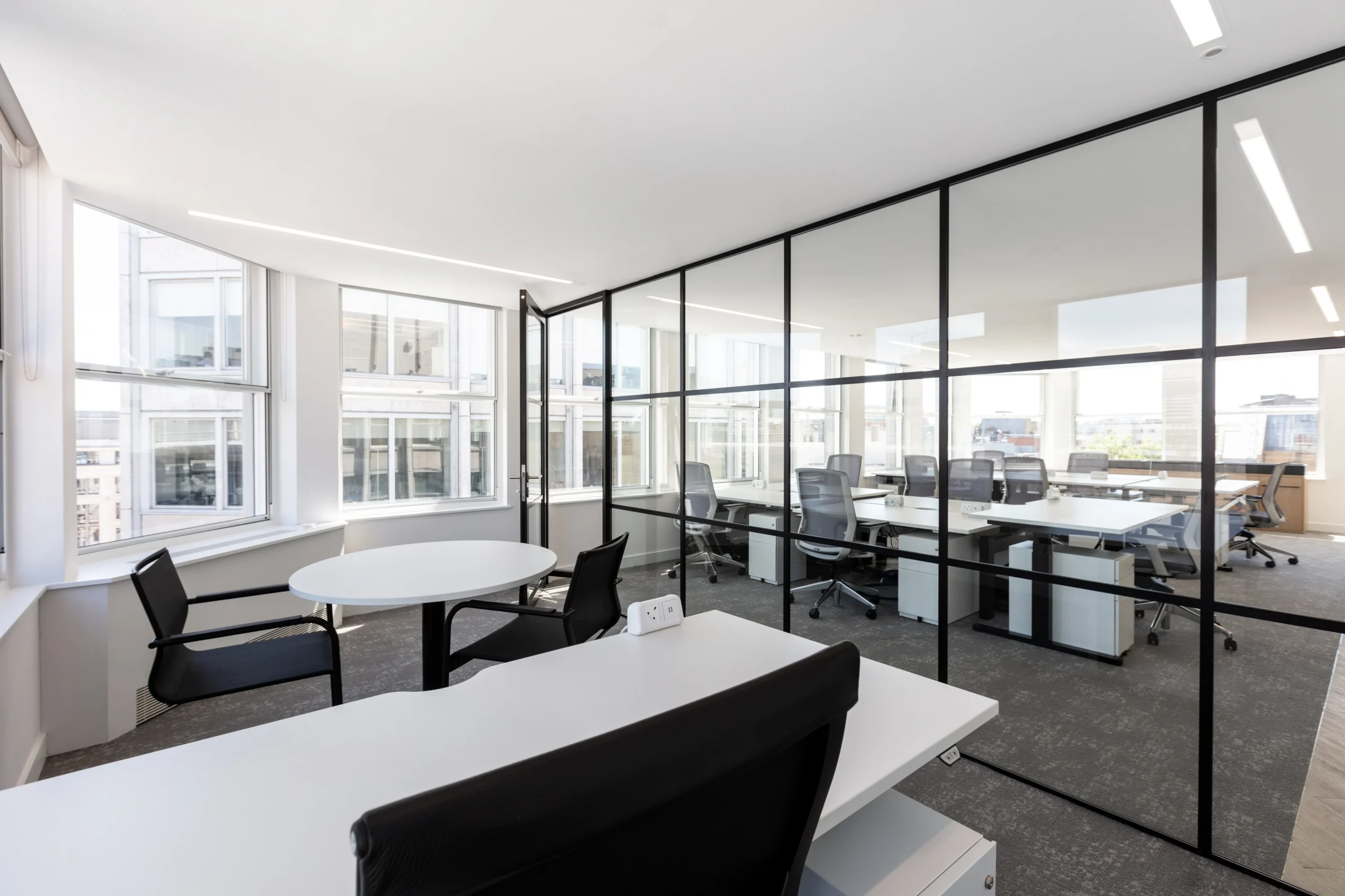 An open-plan office area with floor-to-ceiling glass partitions separating different workspaces. The space features modern furniture, ergonomic chairs, and large windows providing plenty of daylight.