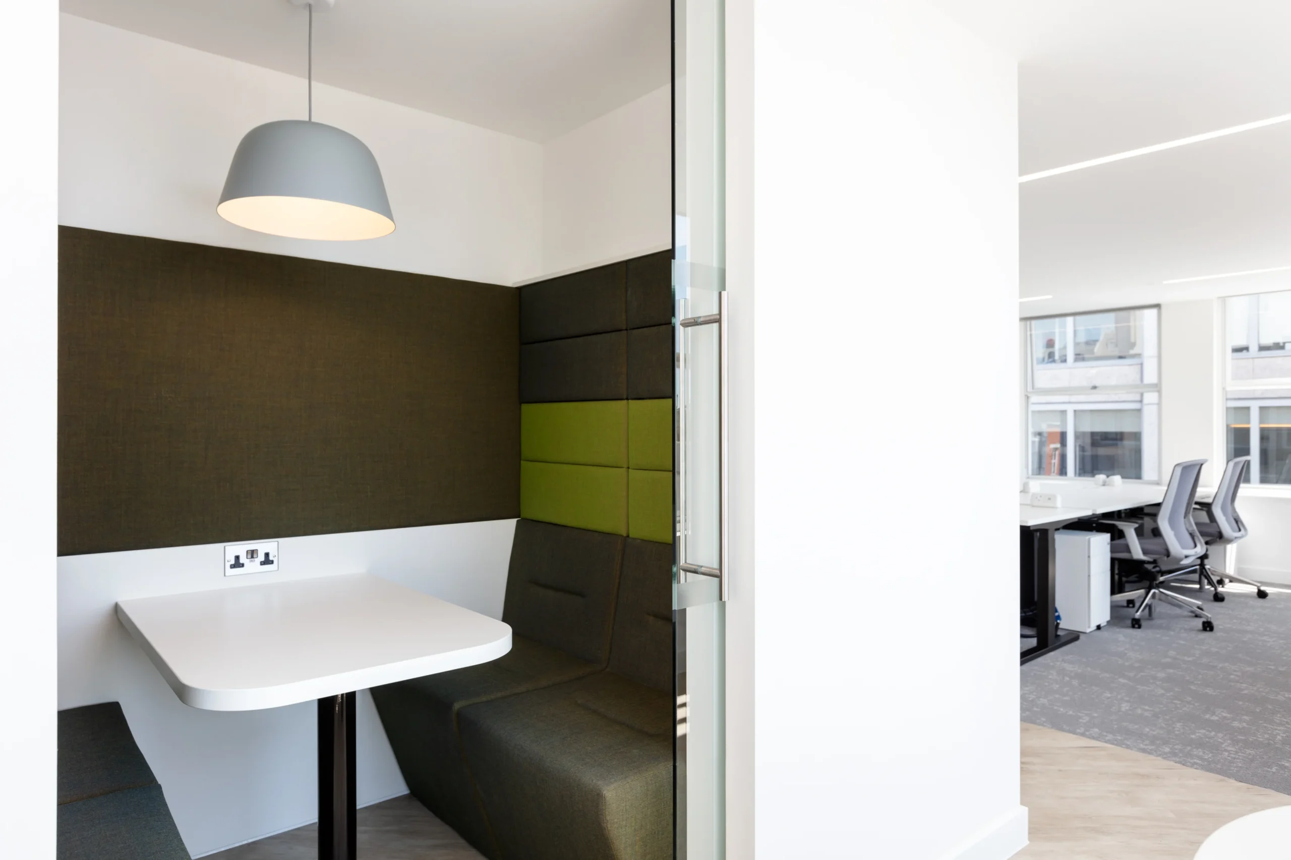 A cosy breakout area with a small table and upholstered bench seating. The area is partitioned with soft green and grey panels for privacy, and a pendant light hangs above for ambient lighting.