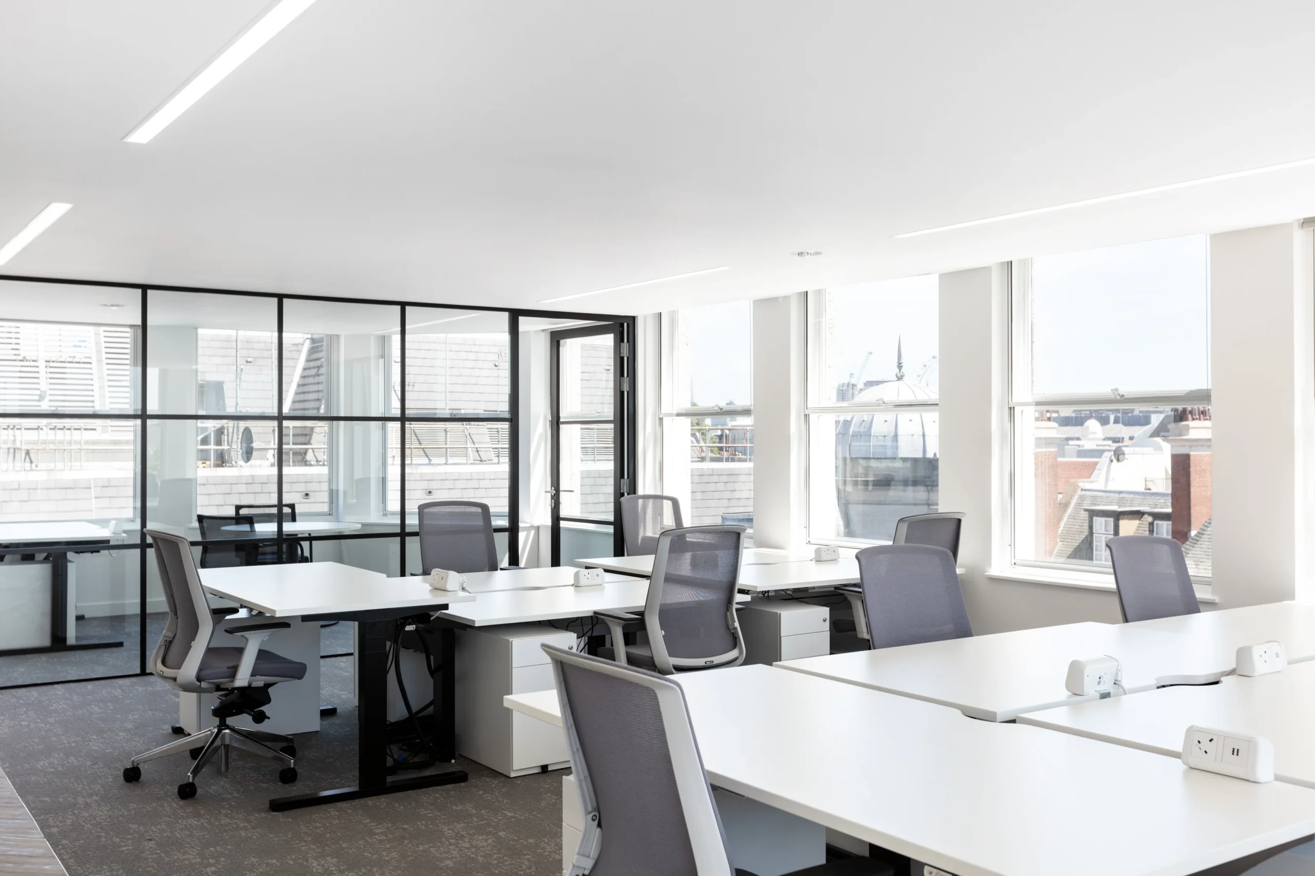 A modern open-plan office setup with several desks and ergonomic chairs. Large windows surround the space.