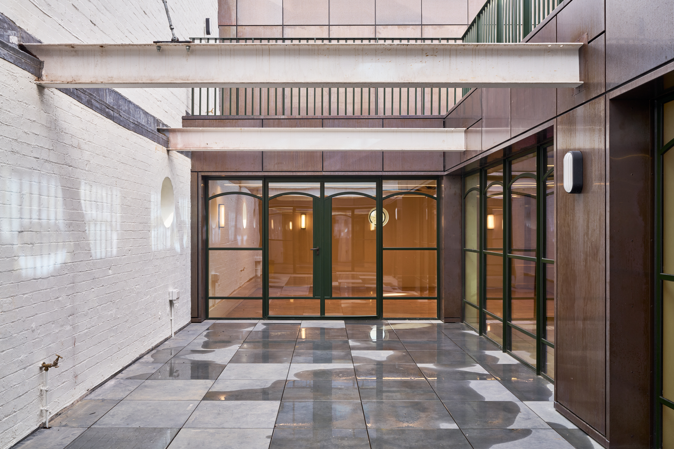 A clean, sleek courtyard space with glass doors framed by metal and dark green structures, leading into a modern indoor area.