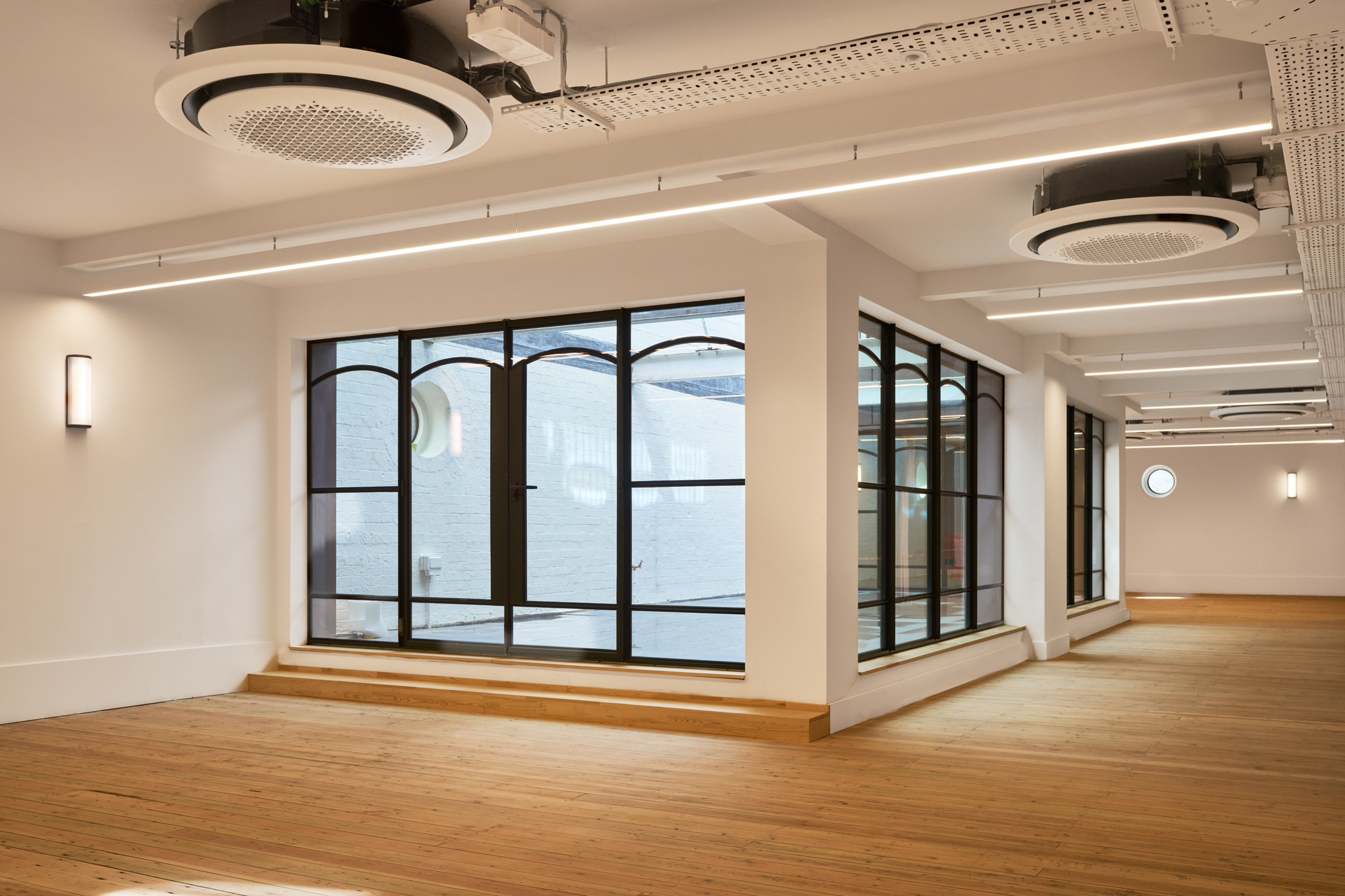 An interior space in Voysey House with large industrial-style windows, clean white walls, and wooden flooring. Small steps lead to an outdoor patio.