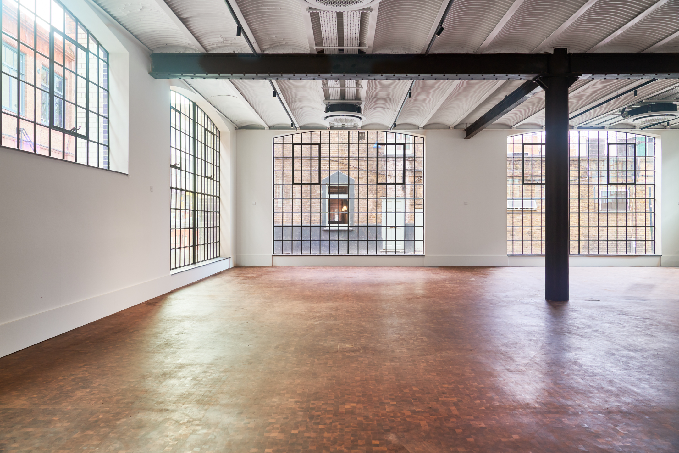 A large industrial-style room with tall, grid-like windows that allow natural light to flood in. The room has a brown polished floor and is framed with exposed steel beams.