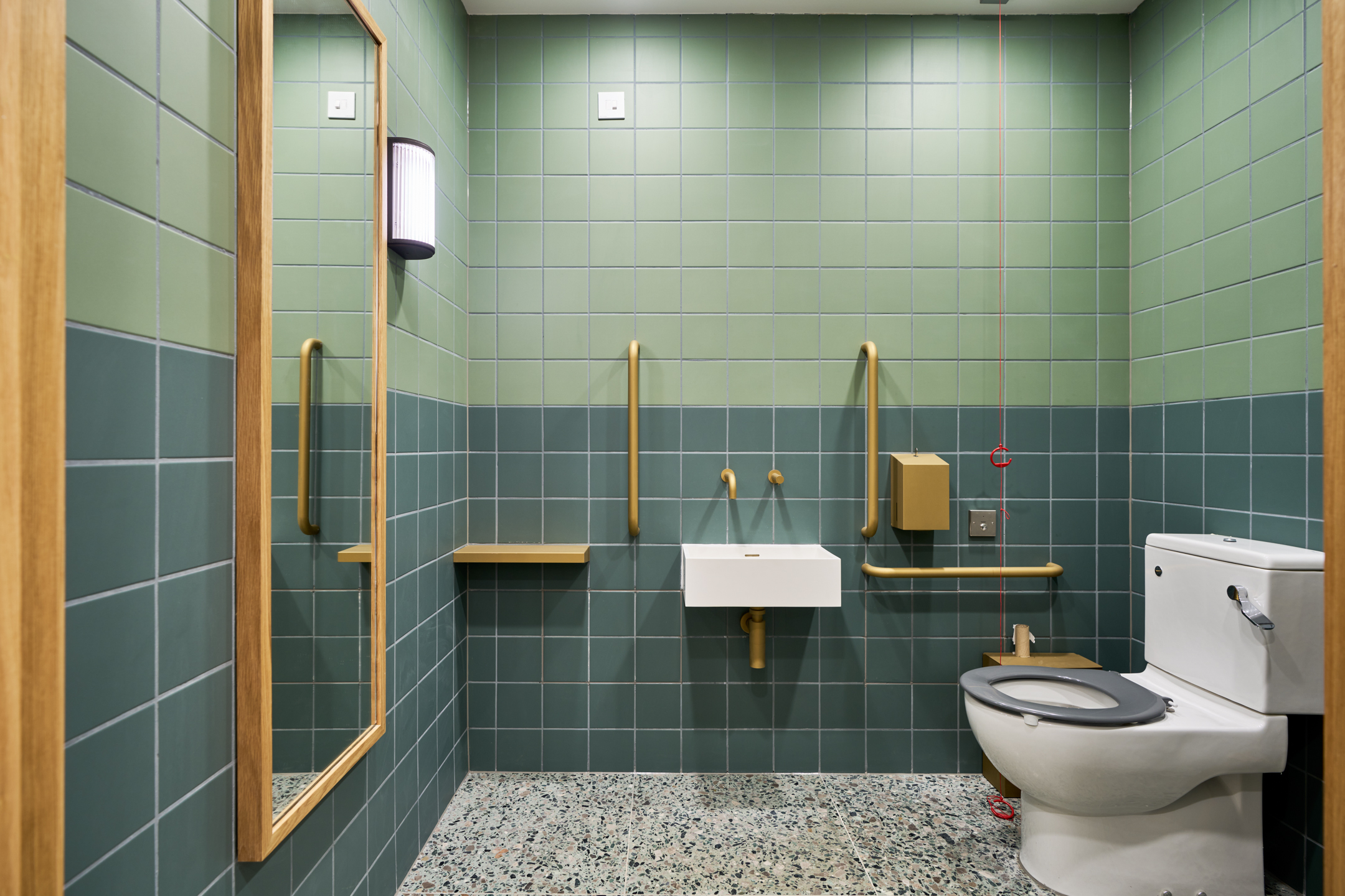 A modern, accessible bathroom at Voysey House with green tiled walls, terrazzo flooring and gold accents on the grab bars, sink, and fixtures.