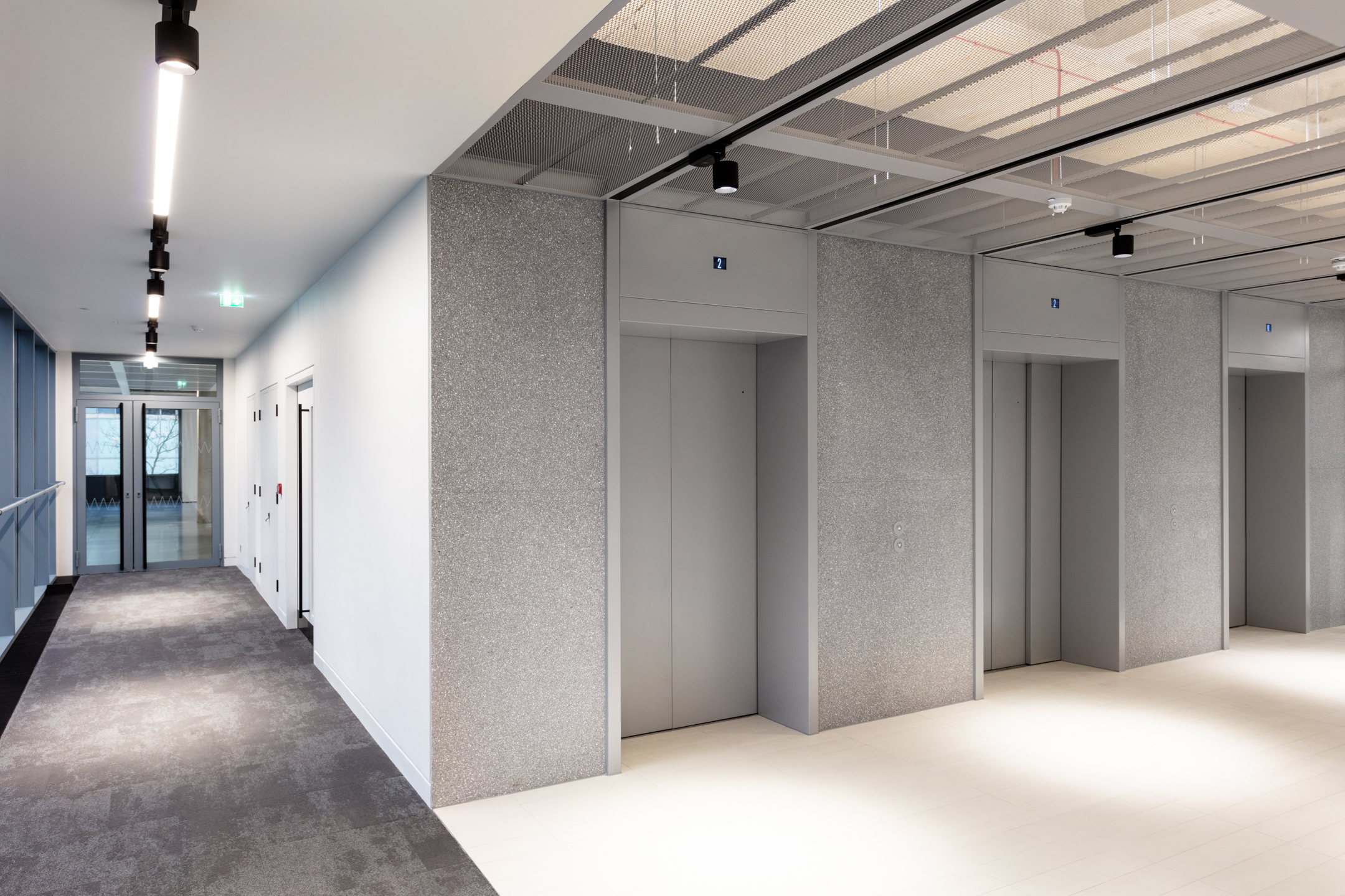 A modern lift lobby at Witan Gate, featuring multiple elevator doors and a minimalist hallway design with a mix of carpet and tile flooring.