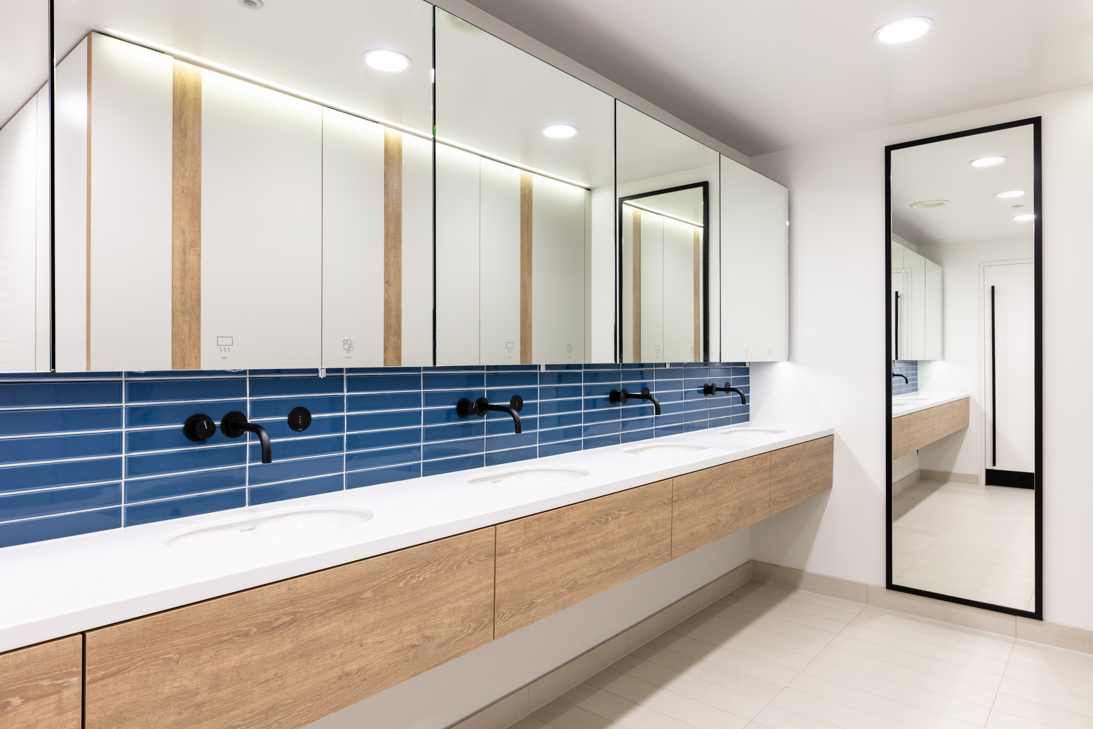 A bathroom at Witan Gate with blue-tiled walls, wooden cabinets, and white countertops. Large mirrors and minimalist lighting give the space a contemporary look.