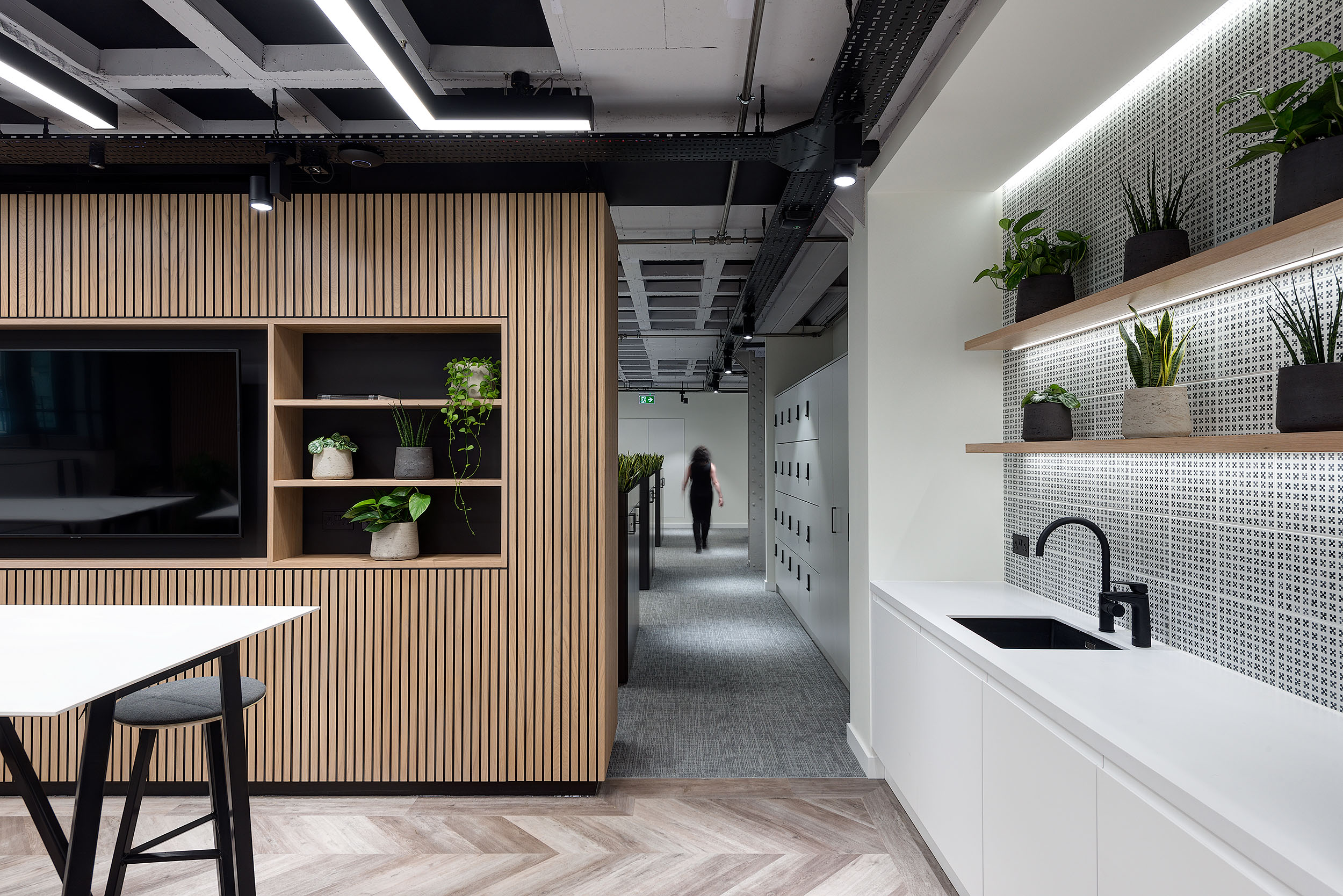 Narrow hallway with a person walking in the background. A white teapoint with black accents and brown tiled flooring are shown.